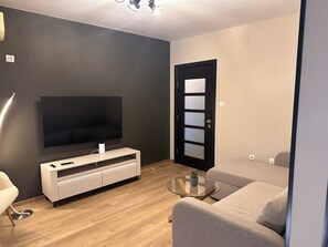 Una sala de estar moderna con un televisor de pantalla plana, un mueble blanco para entretenimiento, una mesa de centro de vidrio y un sofá color beige.
