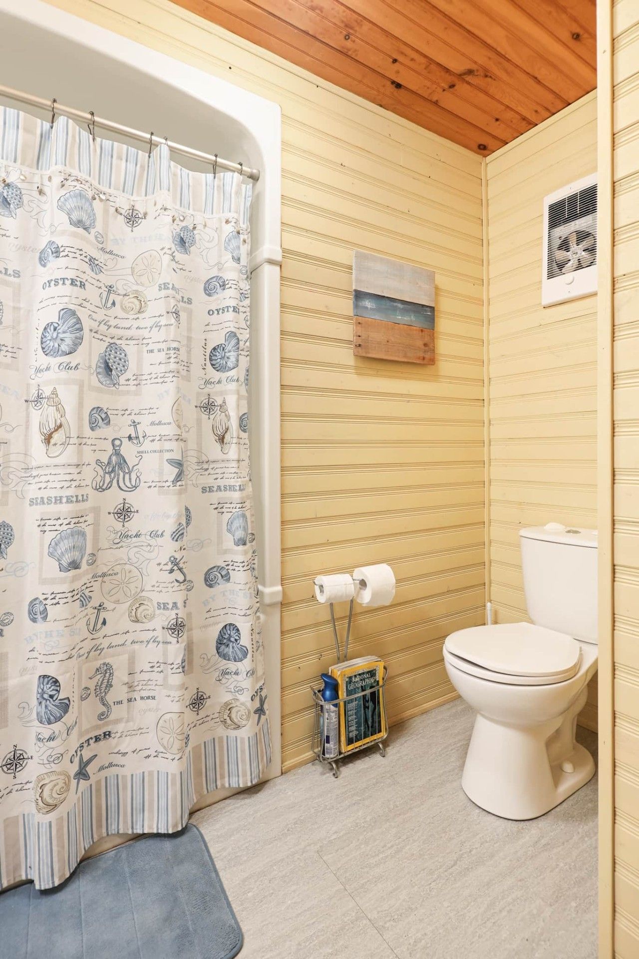 A bathroom with a nautical-themed shower curtain, a toilet, and a small shelf with a magazine.