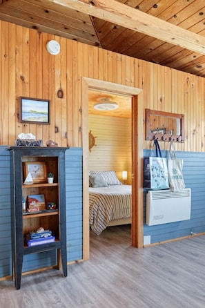 A wooden cabin interior with a bed, a shelf with items, and a wall-mounted clock.