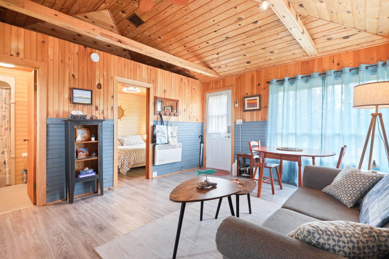 A cozy cabin interior with a wooden ceiling, a dining area, a sofa, and a bed with a striped blanket.