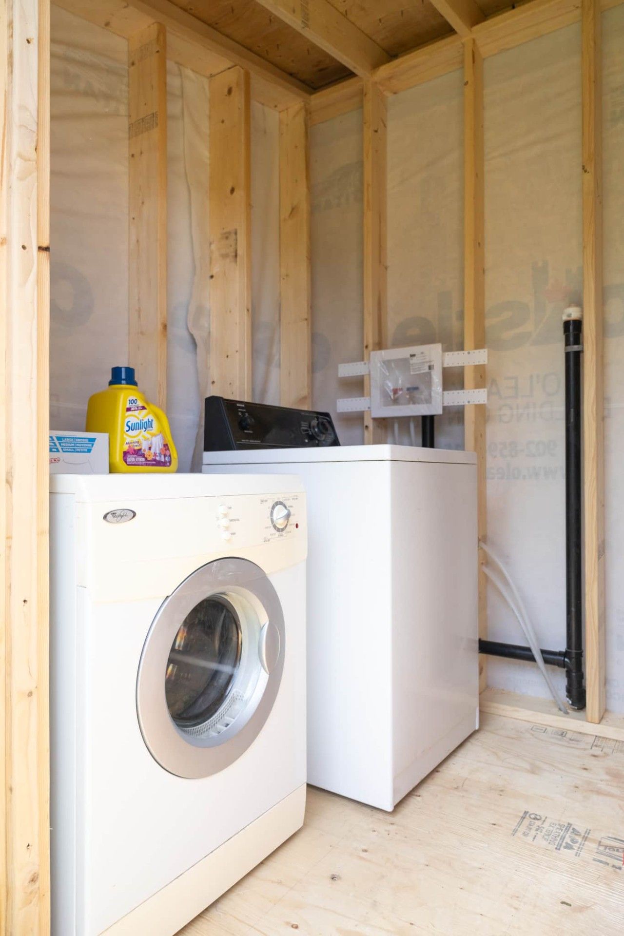 A compact laundry area with a washing machine and dryer, wooden walls, and construction materials visible.