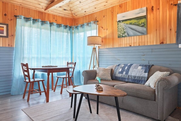 A cozy room with a wooden ceiling, a sofa with patterned cushions, a small table, and a dining table with chairs.