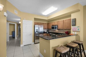 A kitchen with wooden cabinets, a stainless steel refrigerator, a microwave, and a granite countertop island with bar stools. - Sunrise Beach #1609 (Panama City Beach)