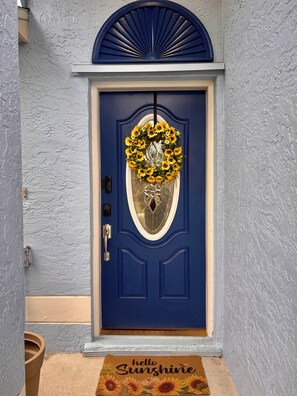Una porta blava amb un disseny de raigs de sol, un panell de vidre amb una corona floral i un estora amb la frase 'hello Sunshine'.
