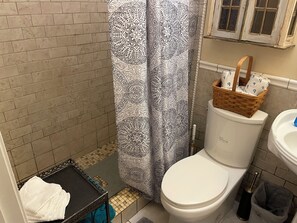 A bathroom with a white toilet, a patterned shower curtain, a wooden basket on the toilet, and a trash bin. - Peaceful Country Atmosphere with City Convenience. (Owens Cross Roads)