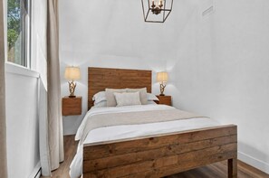 Ein Schlafzimmer mit einem hölzernen Bett, zwei Nachttischen mit Lampen, einem Fenster mit Vorhängen und einer Wandleuchte.