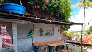 Uma área de estar rústica ao ar livre, com um banco e mesa de madeira, rodeada por árvores e um reservatório azul.
