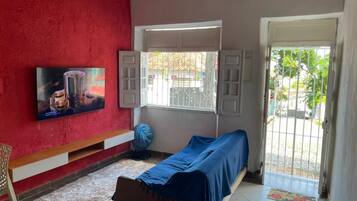 Quarto com cama, televisão fixada numa parede vermelha, janela com vista para o exterior e porta que dá para uma varanda.