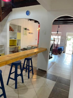 A modern interior with a wooden dining table, blue stools, a white wall with shelves, and a hanging chair.