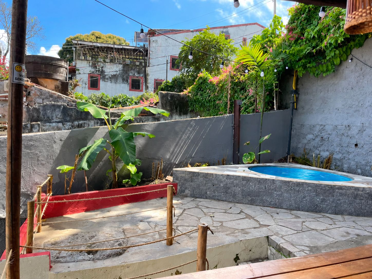 Una piscina con un deck de piedra, rodeada de follaje y una pared gris.