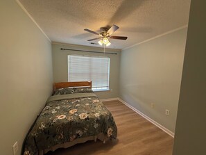 Une chambre à coucher avec un lit, un ventilateur de plafond, une fenêtre avec des stores et un plancher en bois.
