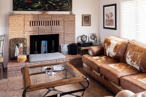 A living room with a brick fireplace, a leather sofa, a coffee table, and a painting on the wall. - 3BR Desert Retreat: Pool, Deck, Games, Cabin & More w/ Private Hiking: 40 Acres (Joshua Tree)