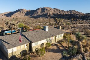 A residential building with a swimming pool, flag, and desert vegetation. - 3BR Desert Retreat: Pool, Deck, Games, Cabin & More w/ Private Hiking: 40 Acres (Joshua Tree)