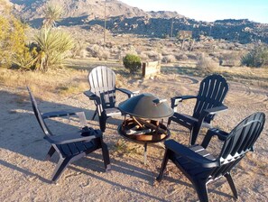 A group of Adirondack chairs arranged around a round table with a grill in a desert landscape. - 3BR Desert Retreat: Pool, Deck, Games, Cabin & More w/ Private Hiking: 40 Acres (Joshua Tree)