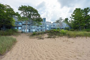 A beachfront property with a modern multi-story building, sandy beach, and surrounding greenery. - Lake Michigan Beachfront | Fireplace + Lake Views (Glen Arbor)