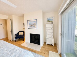 A bedroom with a fireplace, a bed, a chair, a dresser, and a door leading to another room. - Lake Michigan Beachfront | Fireplace + Lake Views (Glen Arbor)