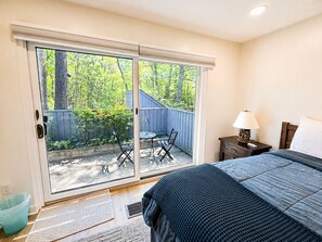A bedroom with a bed, a nightstand, a lamp, and a view through a sliding glass door to an outdoor patio with a table and chairs. - Lake Michigan Beachfront | Fireplace + Lake Views (Glen Arbor)