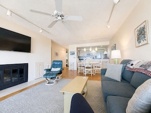 A modern living room with a fireplace, a ceiling fan, a dining area, and a kitchen in the background. - Lake Michigan Beachfront | Fireplace + Lake Views (Glen Arbor)