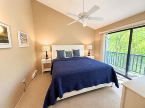 A bedroom with a bed, nightstands, a ceiling fan, and a window with a view of greenery. - Glen Arbor Waterfront | Lake Views + Fireplace (Glen Arbor)