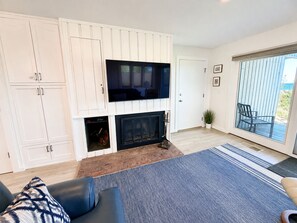 A living room with a flat-screen TV mounted above a fireplace, a sofa, and a large window with a view. - Lake Michigan Beachfront | Private Deck + King Bed (Glen Arbor)