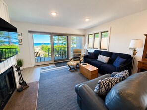A living room with a sofa, armchair, and a view of the beach through large windows. - Lake Michigan Beachfront | Private Deck + King Bed (Glen Arbor)
