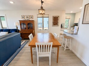 A dining area with a wooden table and white chairs, a blue sofa, and a wooden cabinet with glass doors. - Lake Michigan Beachfront | Private Deck + King Bed (Glen Arbor)