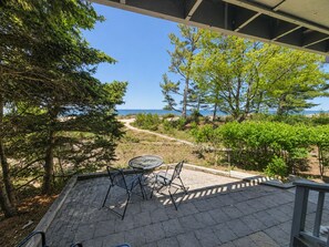 A patio with a table and chairs overlooking a coastal landscape with trees and a clear sky. - Lake Michigan Beachfront | Private Deck + King Bed (Glen Arbor)