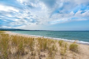 A beach with sand dunes and grass in the foreground, a clear sky, and a calm sea. - Lake Michigan Beachfront | Private Deck + King Bed (Glen Arbor)