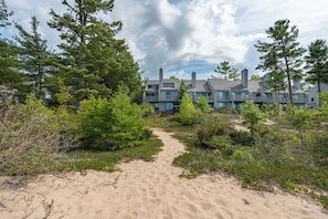 A beachfront property with a sandy path leading to a multi-story building with balconies. - Lake Michigan Beachfront | Private Deck + King Bed (Glen Arbor)