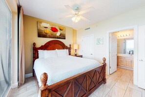 A hotel room with a large bed, a ceiling fan, a painting above the bed, and a bathroom visible through an open door. - Ocean Reef 1502- Blessed Breeze (Panama City Beach)