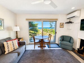 A living room with a sofa, a dining table, and chairs, a fireplace, and a view of the ocean through a sliding glass door. - Beachfront Condo | Lake Views + King Bed (Glen Arbor)