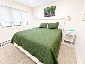 Ein Schlafzimmer mit einem Bett, einem Nachttisch, einer Lampe, einem Fenster mit Jalousien und einem Bild an der Wand.