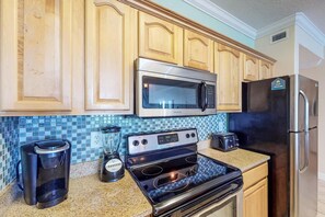 A kitchen with wooden cabinets, a stainless steel microwave, a black refrigerator, a stove, a toaster, and a coffee maker. - Ocean Reef 1703- Ocean O'peace (Panama City Beach)