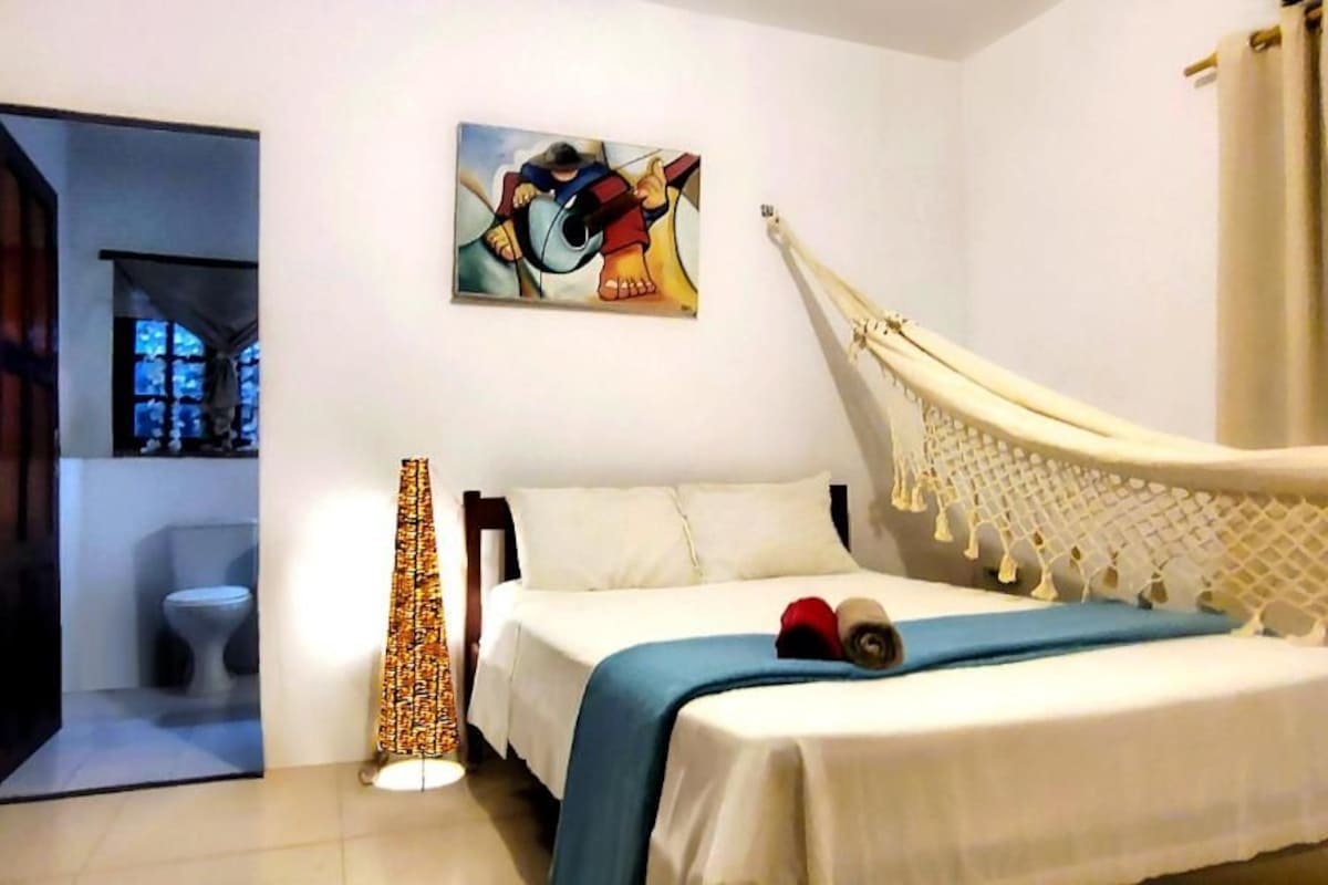 A bedroom with a bed, a hammock, a painting, and a lamp.