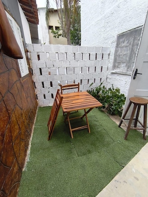Un pequeño patio con una mesa y sillas de madera, rodeado por un muro de ladrillo blanco y un pequeño jardín con plantas.