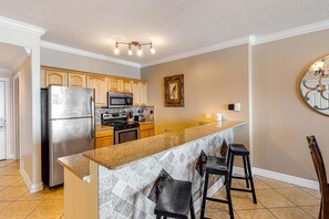 A modern kitchen with a stainless steel refrigerator, a microwave, and a stove. There is a bar area with stools and a decorative wall piece. - Ocean Reef 1702- Gulf of Grace (Panama City Beach)