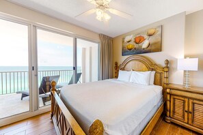 Een hotelkamer met een groot bed, een houten hoofdbord, een bloemschilderij, een nachtkastje en uitzicht op de oceaan door een schuifdeur.