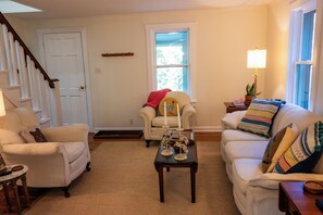 Un salon avec un canapé, un fauteuil et une table basse. On y trouve une porte, une fenêtre et une lampe sur une table d’appoint.
