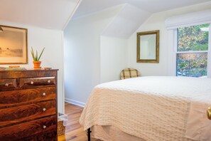 Ein Schlafzimmer mit einem hölzernen Schminktisch, einem Bett mit weißer Bettdecke, einem Fenster mit Blick auf Bäume und einem Spiegel an der Wand.