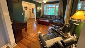 Ein Wohnzimmer mit einem Ecksofa, einem Couchtisch und einem Fernseher. Es gibt einen Deckenventilator, eine Lampe und ein Fenster mit Vorhängen.