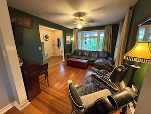 Ein Wohnzimmer mit einem Ecksofa, einem Couchtisch und einem Fernseher. Es gibt einen Deckenventilator, eine Lampe und ein Fenster mit Vorhängen.