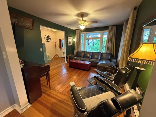 A living room with a sectional sofa, a coffee table, and a TV. There is a ceiling fan, a lamp, and a window with curtains.