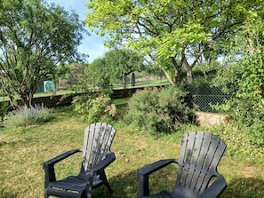 Zwei Adirondack-Stühle in einer grünen Außenanlage mit Bäumen und einem Zaun im Hintergrund.