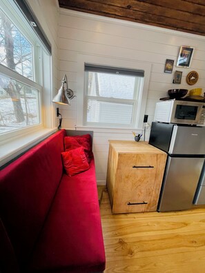 A compact kitchen with a red sofa, wooden cabinets, a microwave, and a window with blinds.