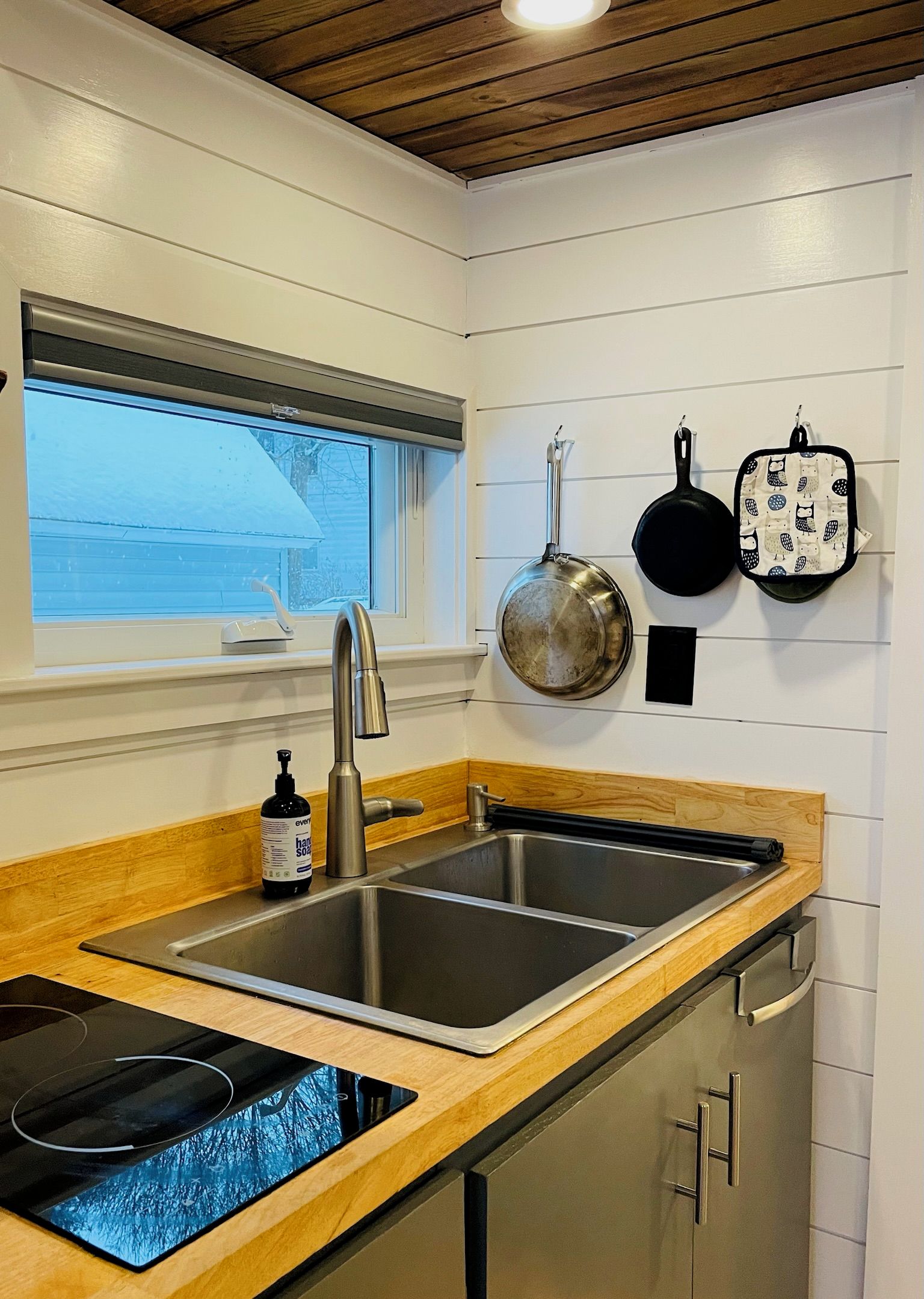 Una cocina con un lavamanos doble, encimera de madera, armarios de acero inoxidable, una ventana con persianas y utensilios de cocina colgantes.