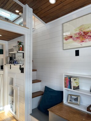 Een kamer met een houten plafond, witte muren, een boekenplank, een ingelijste foto en een raam met uitzicht.