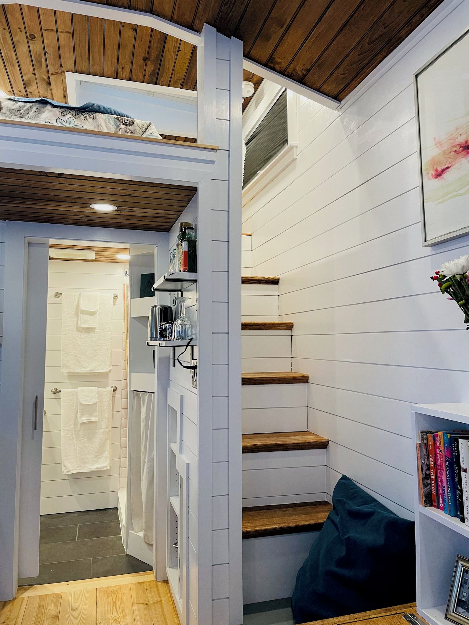 A narrow hallway with a wooden ceiling, white walls, a staircase, and a bookshelf with books.