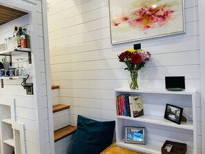 Una habitación con paredes de paneles blancos, una banca de madera, un cojín azul turquesa, un jarrón con flores, un cuadro enmarcado y un estante con libros y un pequeño dispositivo electrónico.