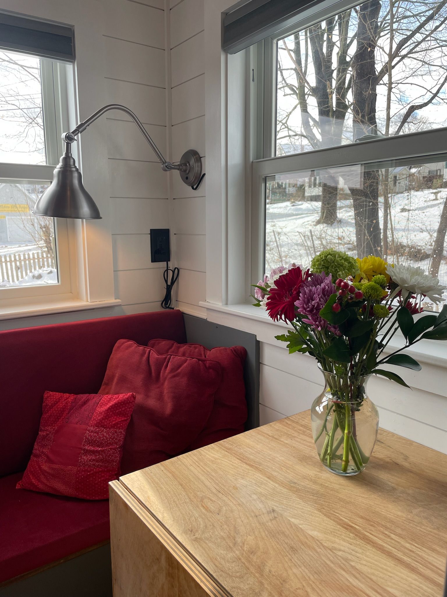 A room with a red sofa, a wooden table, a vase with flowers, and a window with a view of trees and snow.
