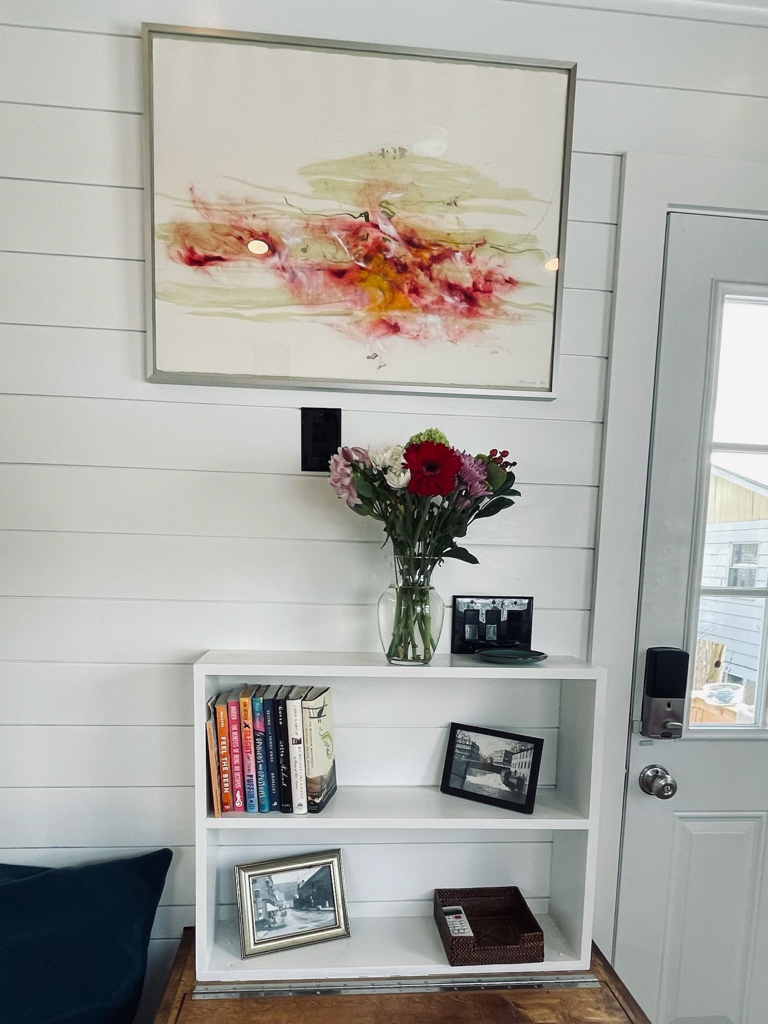 A white wall with a framed abstract painting, a vase with flowers, a bookshelf with books and a framed photo, and a door with a keypad lock.